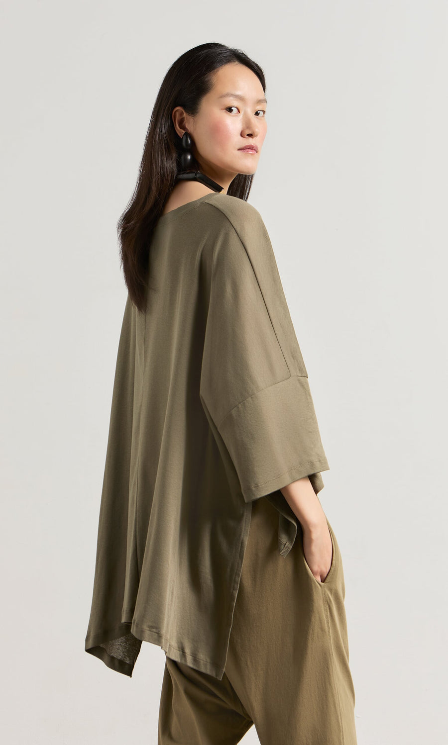 Plümo- Woman in an oversized olive-green Kaia Jersey boatneck top, standing sideways and looking over her shoulder.