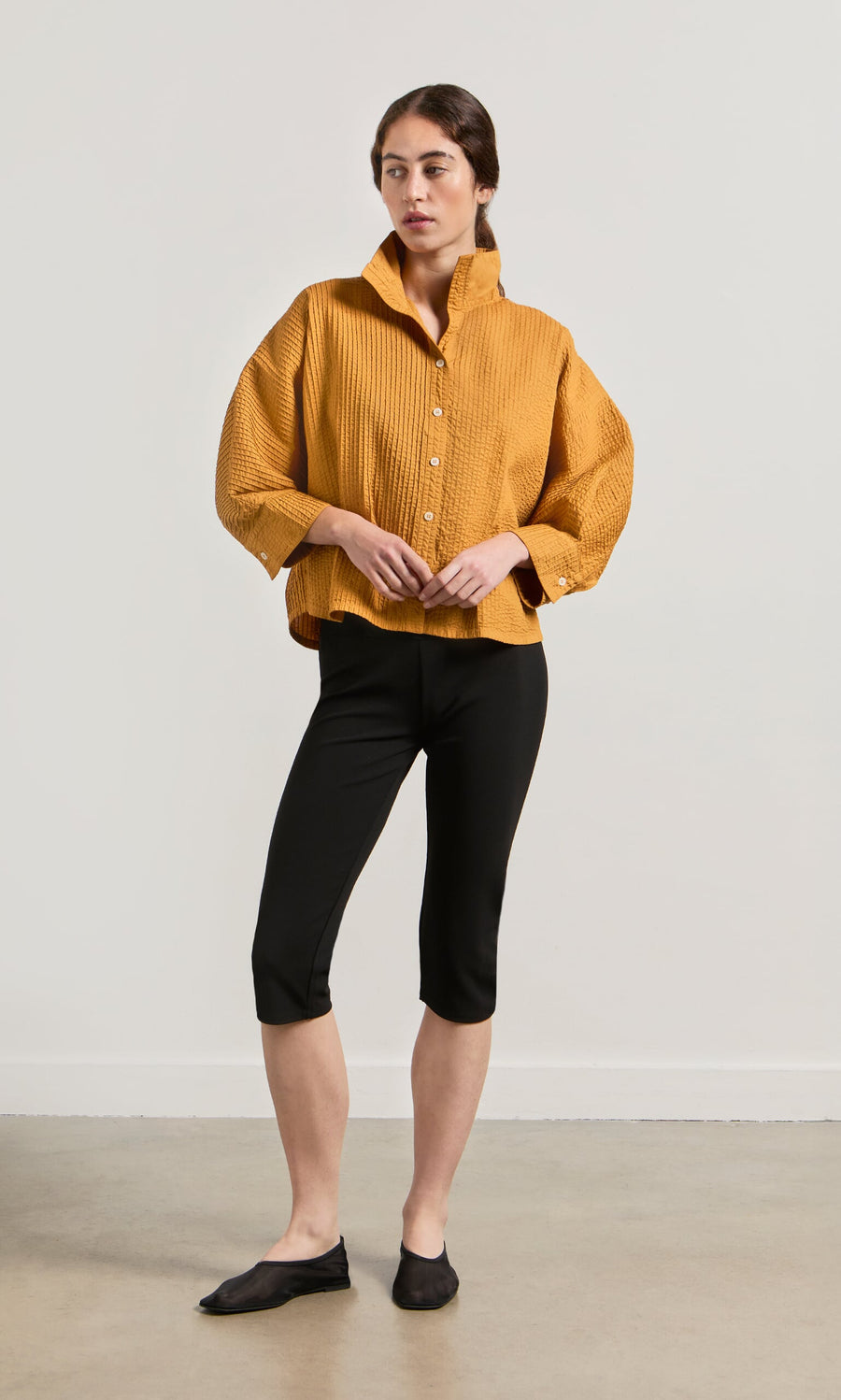 Plümo- Woman wearing a loose mustard yellow Karashi Shirt, black capri pants, and black flats against a plain background.