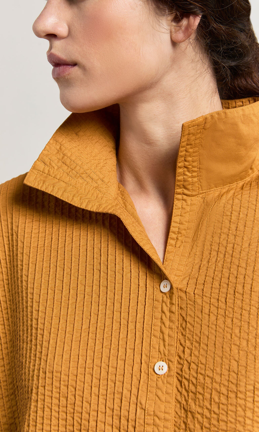 Plümo- A woman wears the Karashi Shirt, a textured mustard pintuck top with a high collar and wooden buttons.