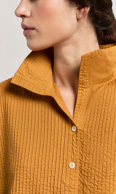 Plümo- A woman wears the Karashi Shirt, a textured mustard pintuck top with a high collar and wooden buttons.