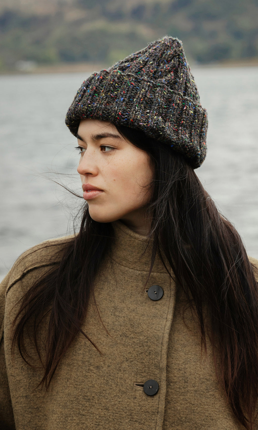 Plümo  Woman in a Kilcarra Tweed Beanie and brown coat stands by water, gazing calmly to the side.