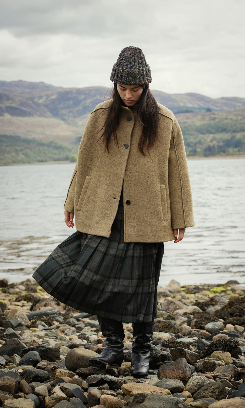 Plümo  Woman in tan coat and plaid skirt, wearing Kilcarra Tweed Beanie, stands by rocky shore with water and hills behind.