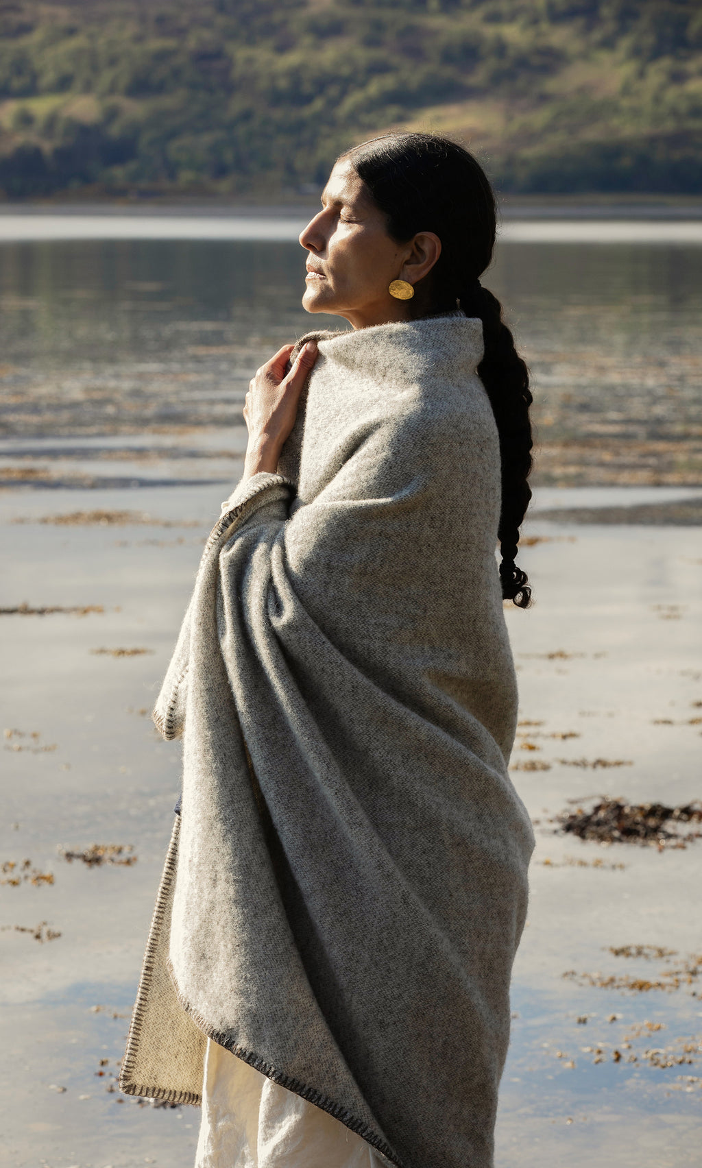 Plümo  Woman with long dark hair in Kilkenny Blanket - Fossil stands by water, eyes closed, basking in the sunlight.