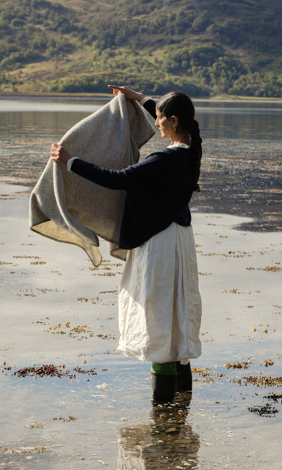 Plümo  A woman stands in shallow water holding the Kilkenny Blanket - Fossil; hills and trees are in the background.