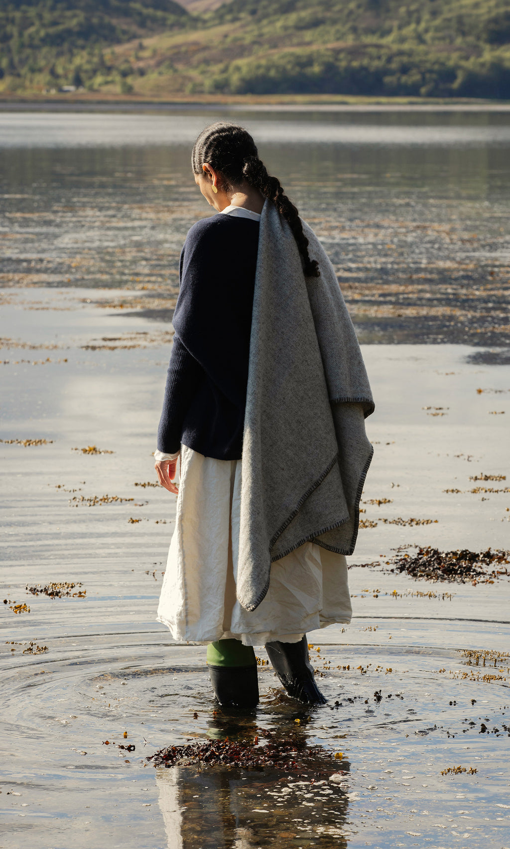 Plümo  Wearing boots, a white skirt, and the Kilkenny Blanket - Fossil, a person stands by the lakeshore facing away.