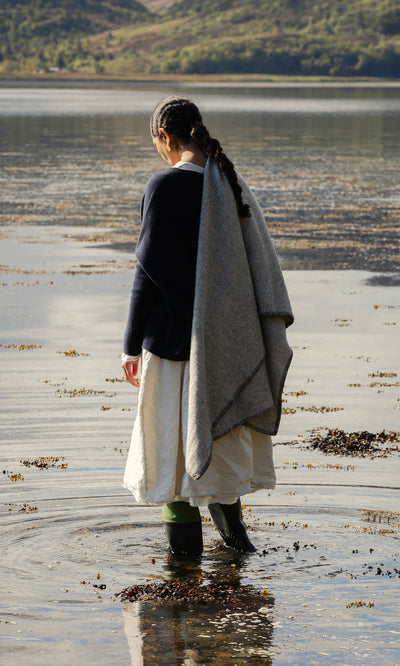 Plümo  Wearing boots, a white skirt, and the Kilkenny Blanket - Fossil, a person stands by the lakeshore facing away.