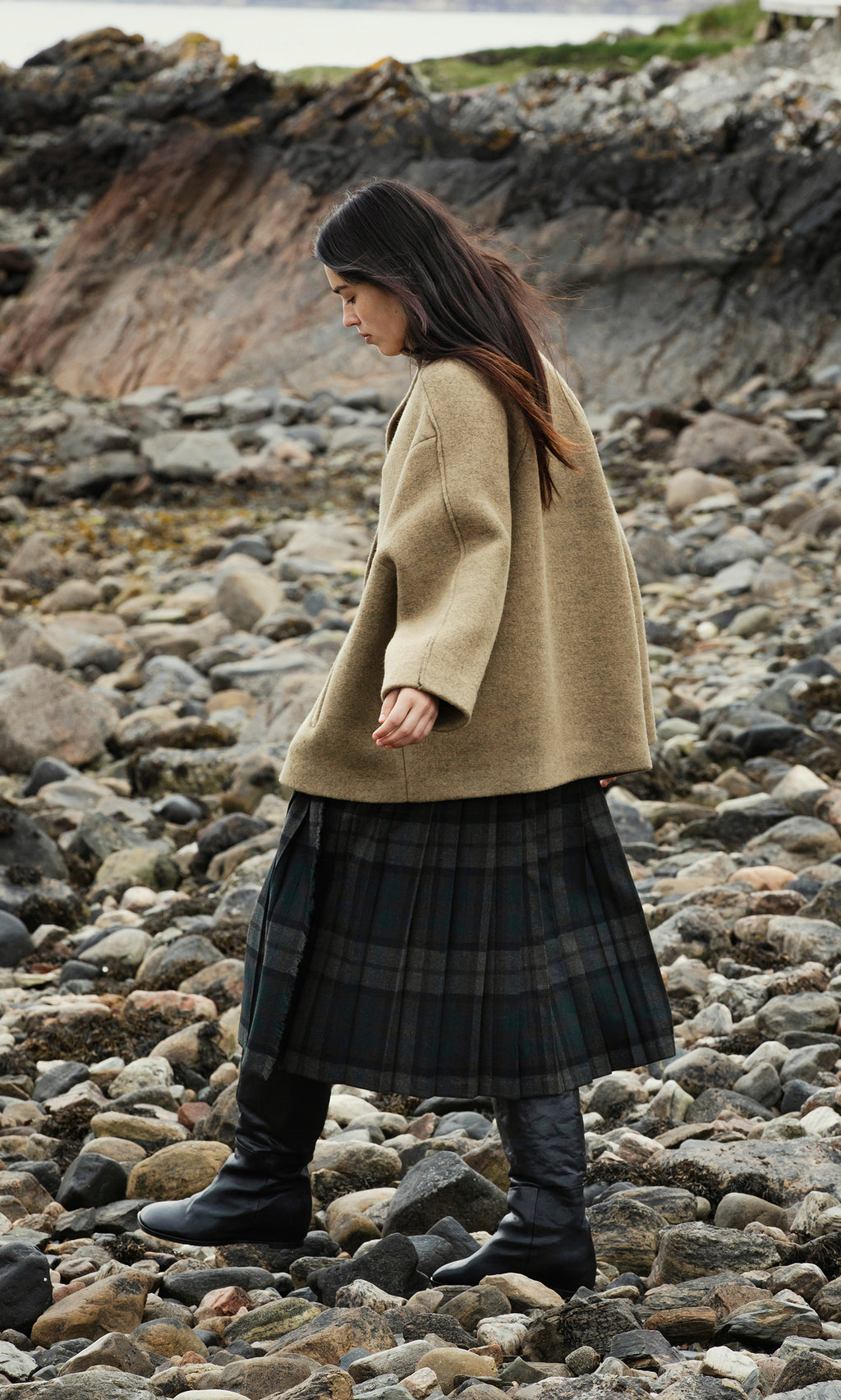 Plümo  A person in the acorn brown Kilmore Coat and plaid skirt walks on a rocky shore with dark rocks behind them.
