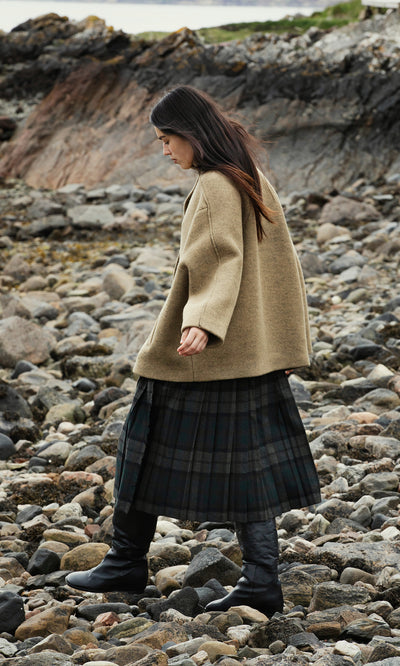 Plümo  Woman in a tan coat and Campbell Kilt walks on a rocky shoreline with cliffs and water behind her.