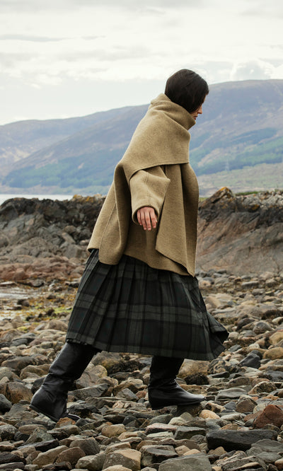 Plümo  Person in tan shawl and Campbell Kilt walks on rocky ground by water and mountains, looking down.