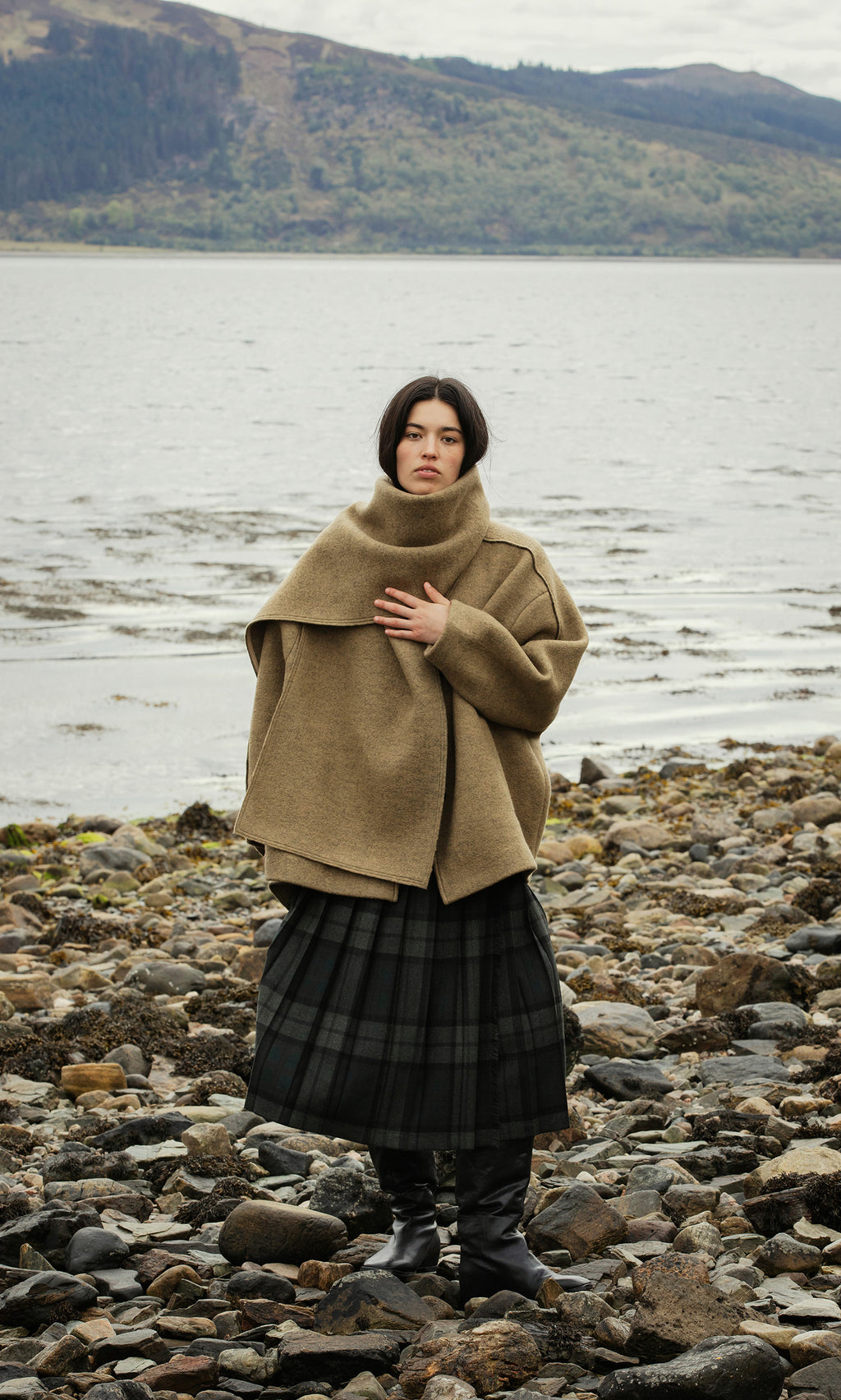 Plümo  A woman in the Kilmore Coat stands on a rocky shore, water and hills behind her, layered in chic plaid.