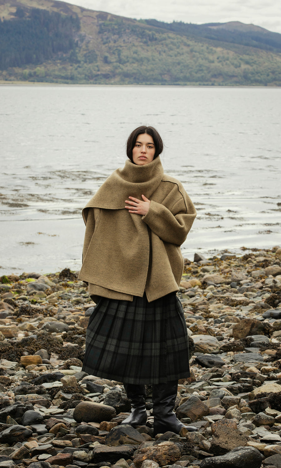 Plümo  A woman in the Kilmore Coat stands on a rocky shore, water and hills behind her, layered in chic plaid.