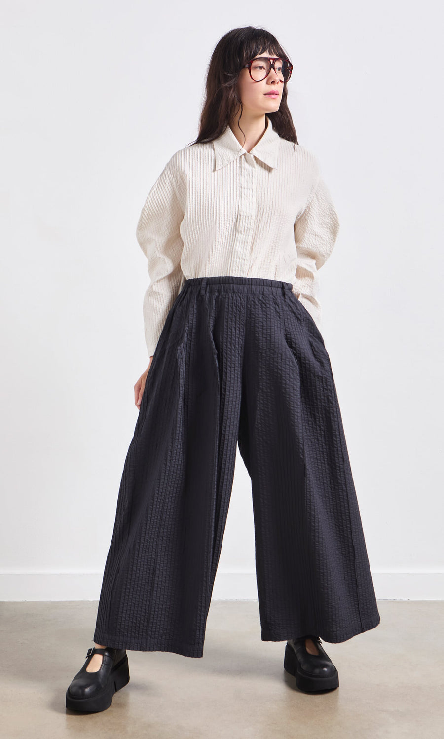 Plümo- Person in a cream textured shirt, glasses, and Kiri Pintuck Pants stands against a plain background.