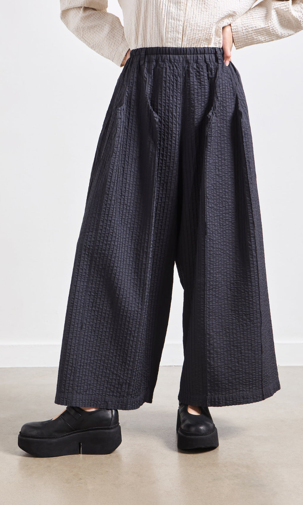 Plümo- Person wearing Kiri Pintuck Pants with vertical pleating and black platform shoes, standing against a white wall.