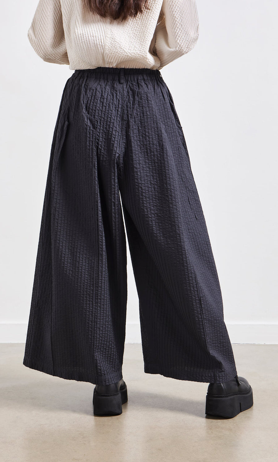 Plümo- Someone wearing Kiri Pintuck Pants with vertical pleats and black platform shoes, viewed from the back.
