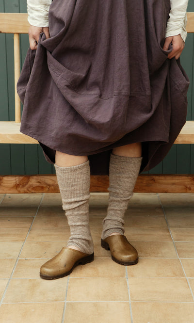 Plümo  Person in Opana Mules, a purple skirt, and beige socks stands on tile floor by a wooden bench.