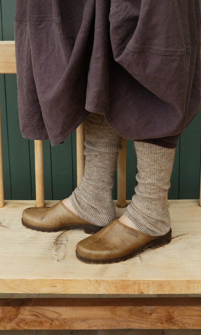 Plümo  Person wearing Opana Mules, a long brown skirt, and thick beige socks stands on a wooden bench.