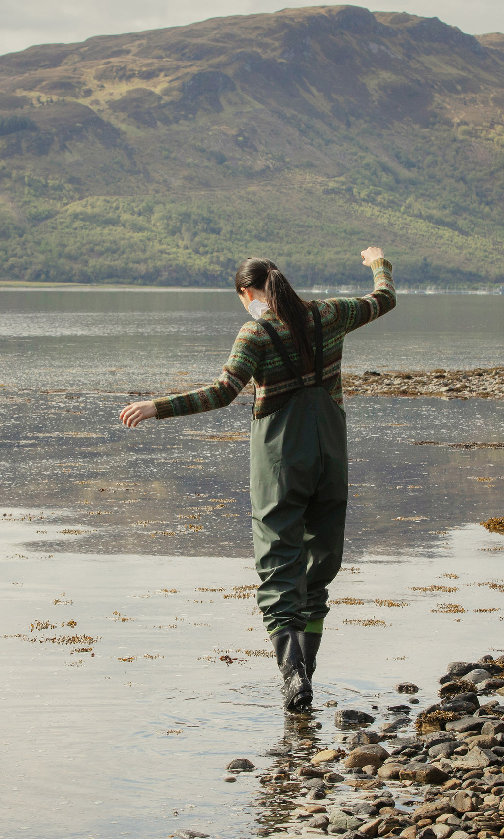 Plümo  Person in Land Dungarees balances on lakeside rocks with mountains, trees, and a cloudy sky in the background.