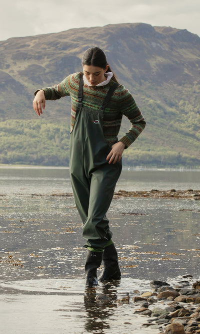 Plümo  Person in Land Dungarees and a sweater walks along a rocky shoreline with hills in the background.