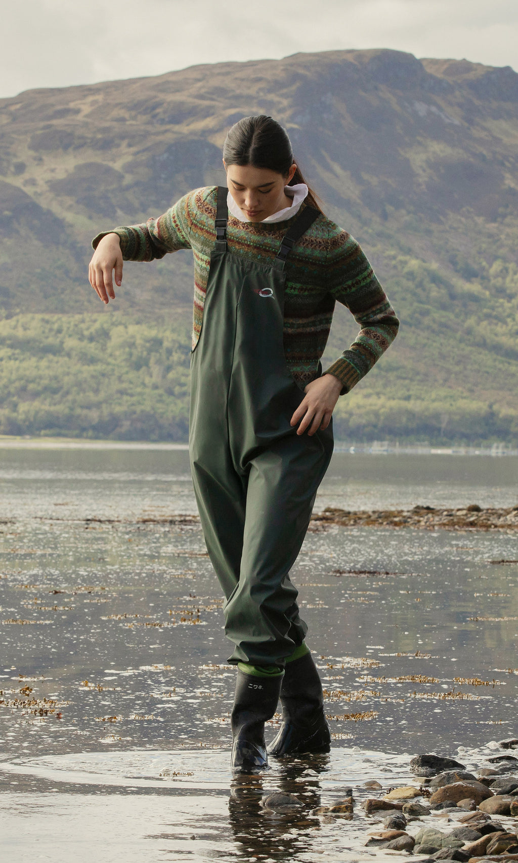 Plümo  Wearing weatherproof Land Dungarees, someone walks in shallow water along a rocky shore with hills in the background.