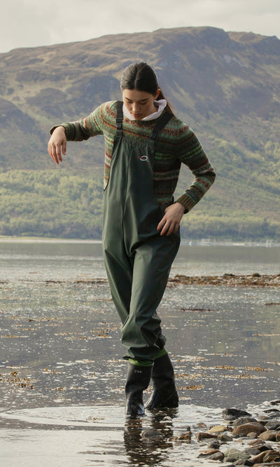 Plümo  Wearing weatherproof Land Dungarees, someone walks in shallow water along a rocky shore with hills in the background.
