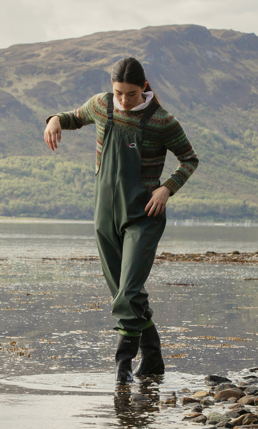 Plümo  Wearing weatherproof Land Dungarees, someone walks in shallow water along a rocky shore with hills in the background.