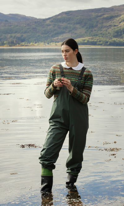 Plümo  A woman in Land Dungarees stands in shallow lake water, with green hills visible in the background.