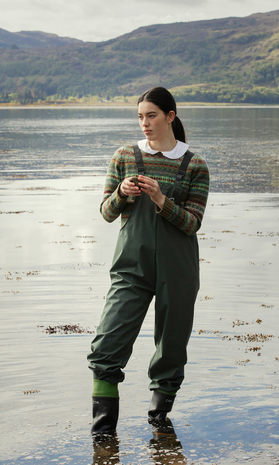 Plümo  A woman in Land Dungarees stands in shallow lake water, with green hills visible in the background.
