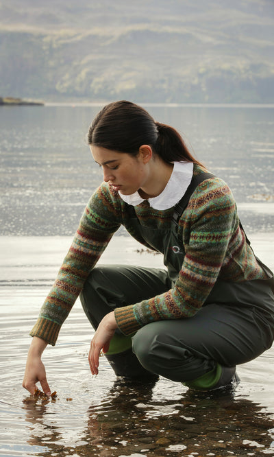 Plümo  A woman in Land Dungarees crouches by a lake, touching the water with her hand on a cloudy day.