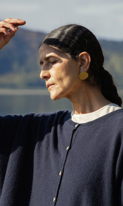 Plümo  Woman with dark hair in a ponytail wears Cohen Earrings and a navy sweater, shading her eyes outside.