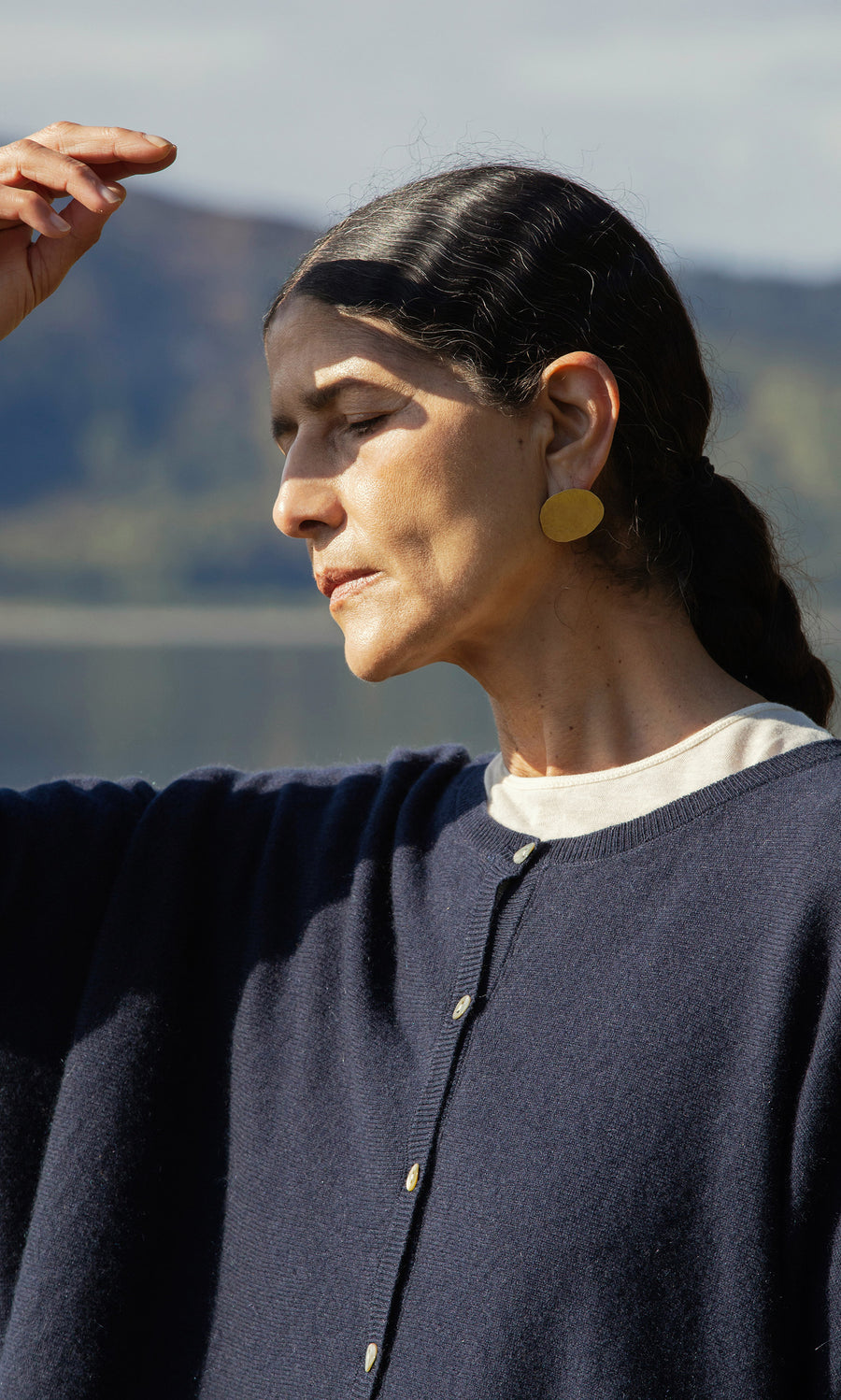 Plümo  Woman with dark hair in a ponytail wears Cohen Earrings and a navy sweater, shading her eyes outside.