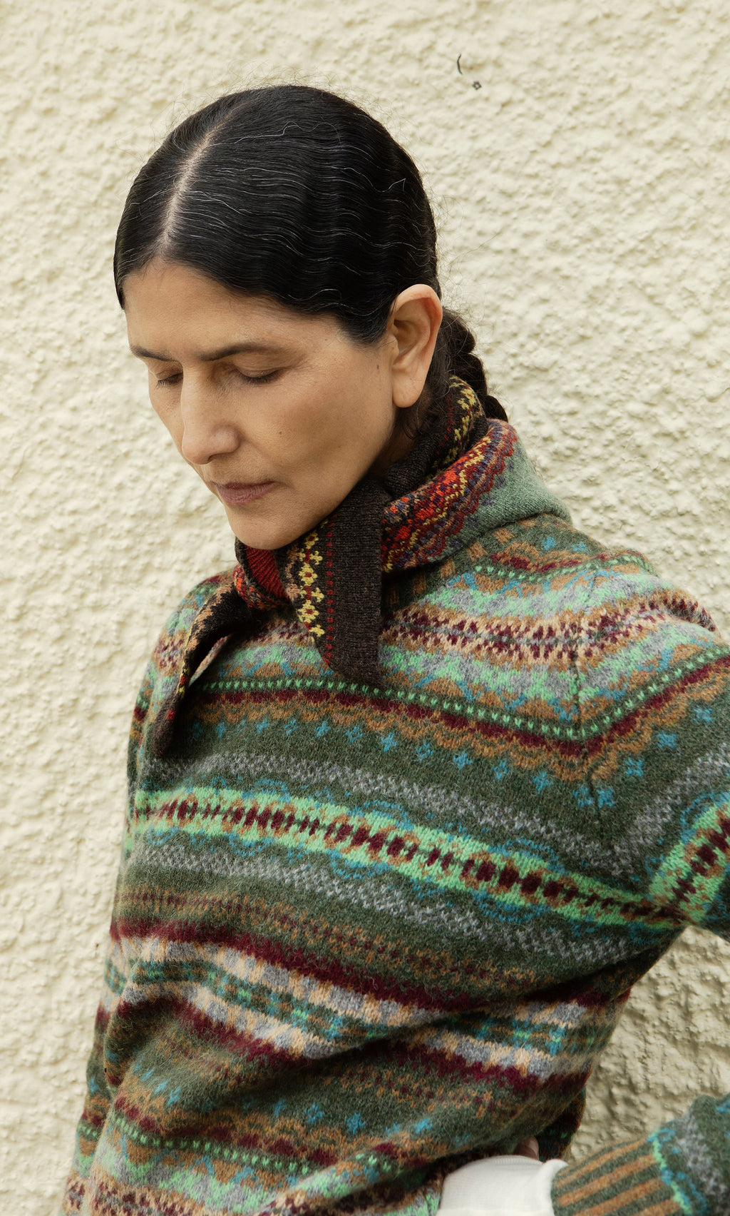 Plümo  Person in pure new wool sweater and Lenie Necktie stands by a textured wall, eyes closed, appearing reflective.