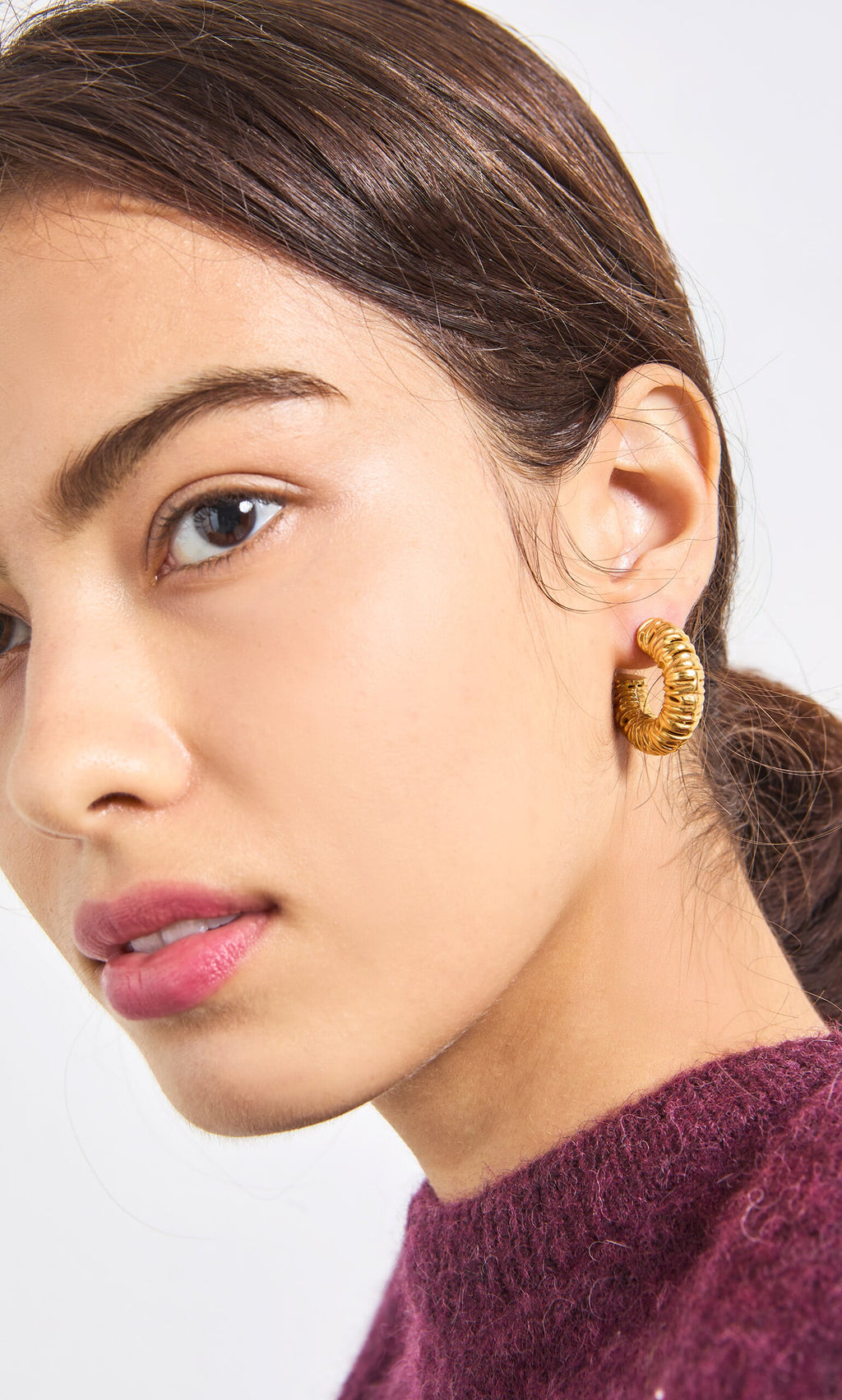 Plümo- Woman with smooth skin wears Lili Hoops gold plated earrings and a maroon sweater, hair tied back, facing sideways.