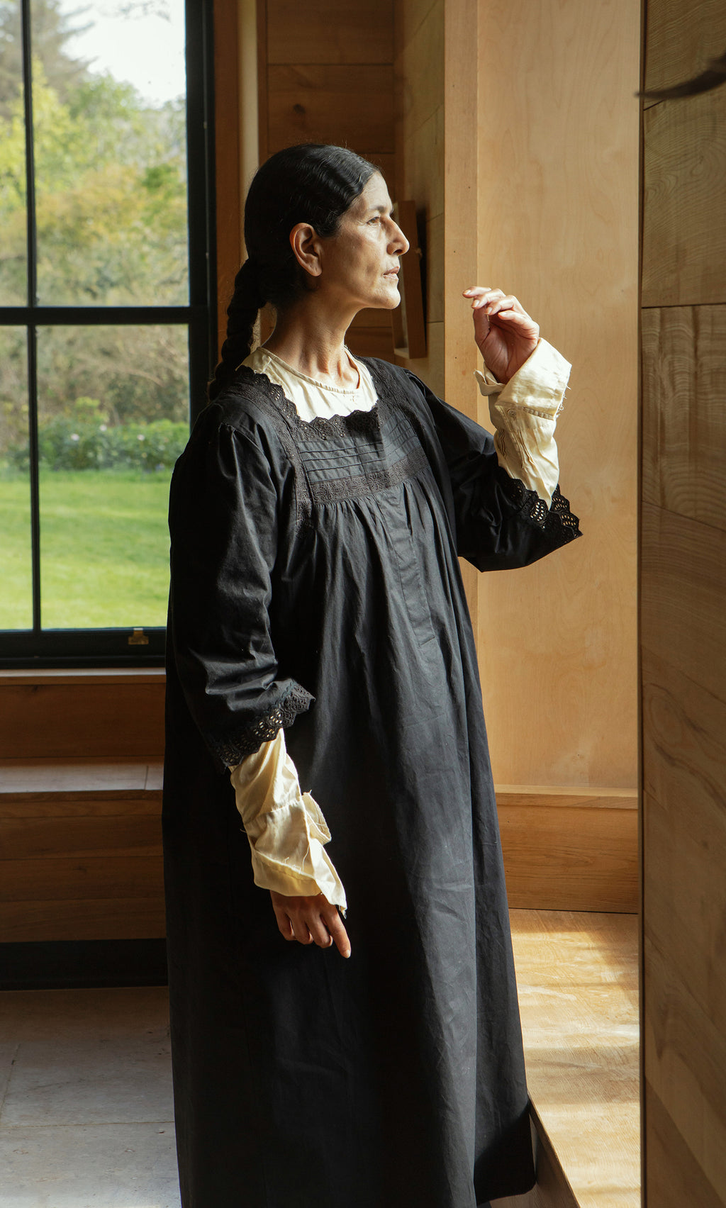 Plümo  A woman in a black Dahlia Dress stands by a window, gazing outside thoughtfully with one hand raised.