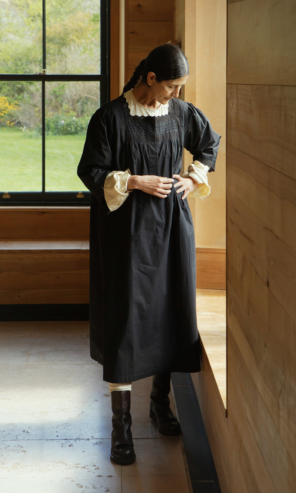 Plümo  A woman in the Dahlia Dress, black cotton with cream ruffles, stands by a window in a wooden room, looking down.