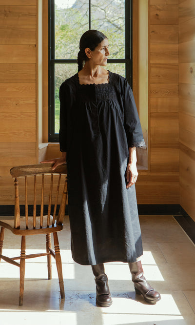 Plümo  Woman in the Dahlia Dress stands by a wooden chair in a sunlit room with wood walls and a window.
