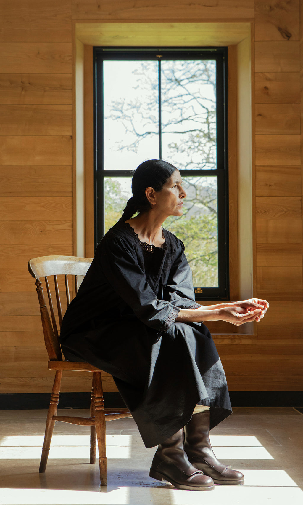 Plümo  A woman in a Dahlia Dress sits on a wooden chair by a window in a sunlit wooden room.
