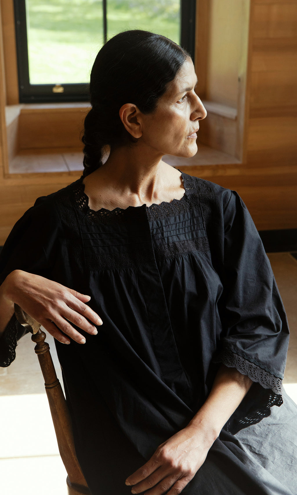 Plümo  A woman in the vintage Dahlia Dress sits on a wooden chair, gazing sideways in a sunlit, wood-paneled room.