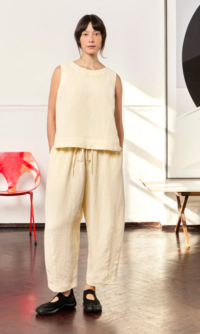 Plümo- Woman indoors wears the Linda Top - Milk, relaxed fit cream linen, with matching wide pants and black shoes. Red chair behind.