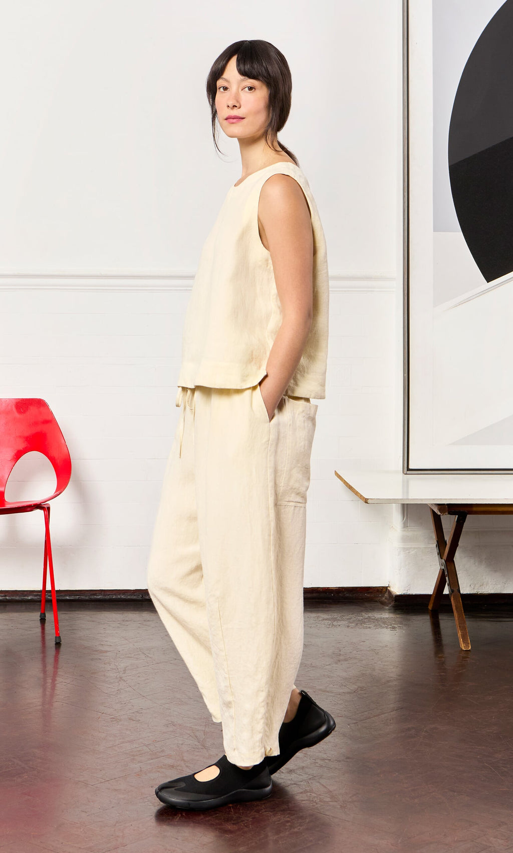 Plümo- Woman in the Linda Top - Milk, hands in pockets, stands indoors beside a red chair and abstract artwork.