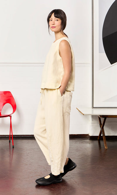 Plümo- Woman in the Linda Top - Milk, hands in pockets, stands indoors beside a red chair and abstract artwork.