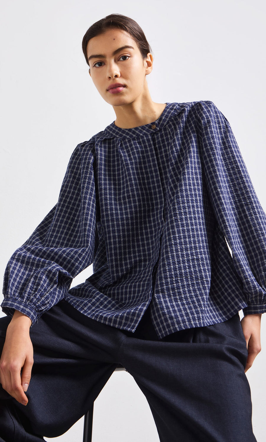 Plümo- Someone wearing the Little Prairie Blouse in blue check and dark pants sits on a stool against a plain background.