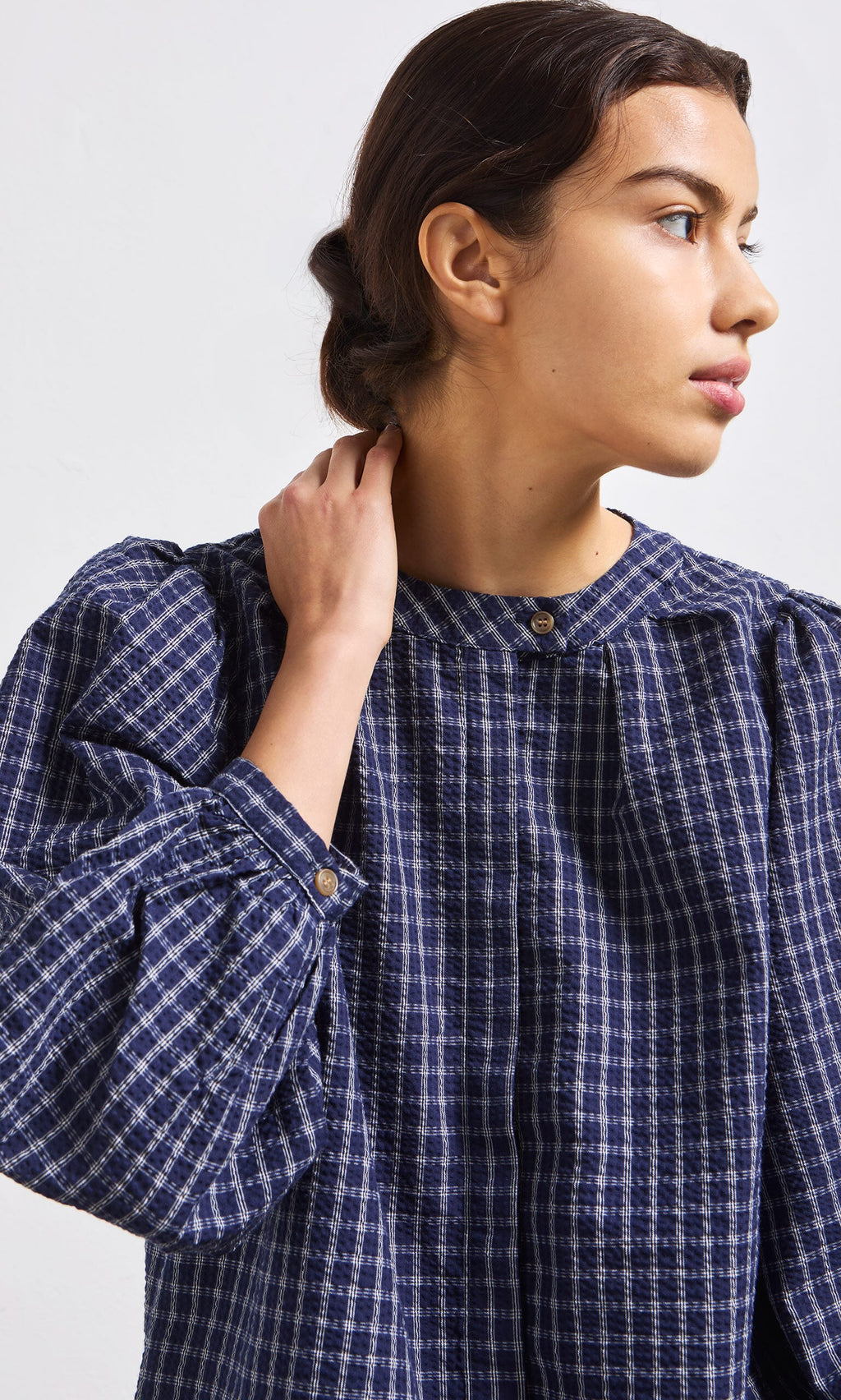 Plümo- Woman in the Little Prairie Blouse, a blue check button-up with puffed sleeves, looks to the side, hand at her neck.