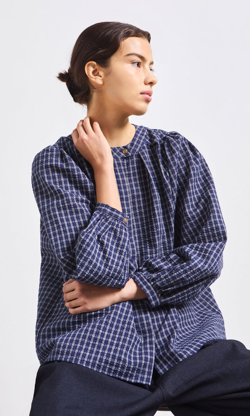 Plümo- A person in a Little Prairie Blouse and dark pants sits with one arm crossed, gazing thoughtfully to the side.
