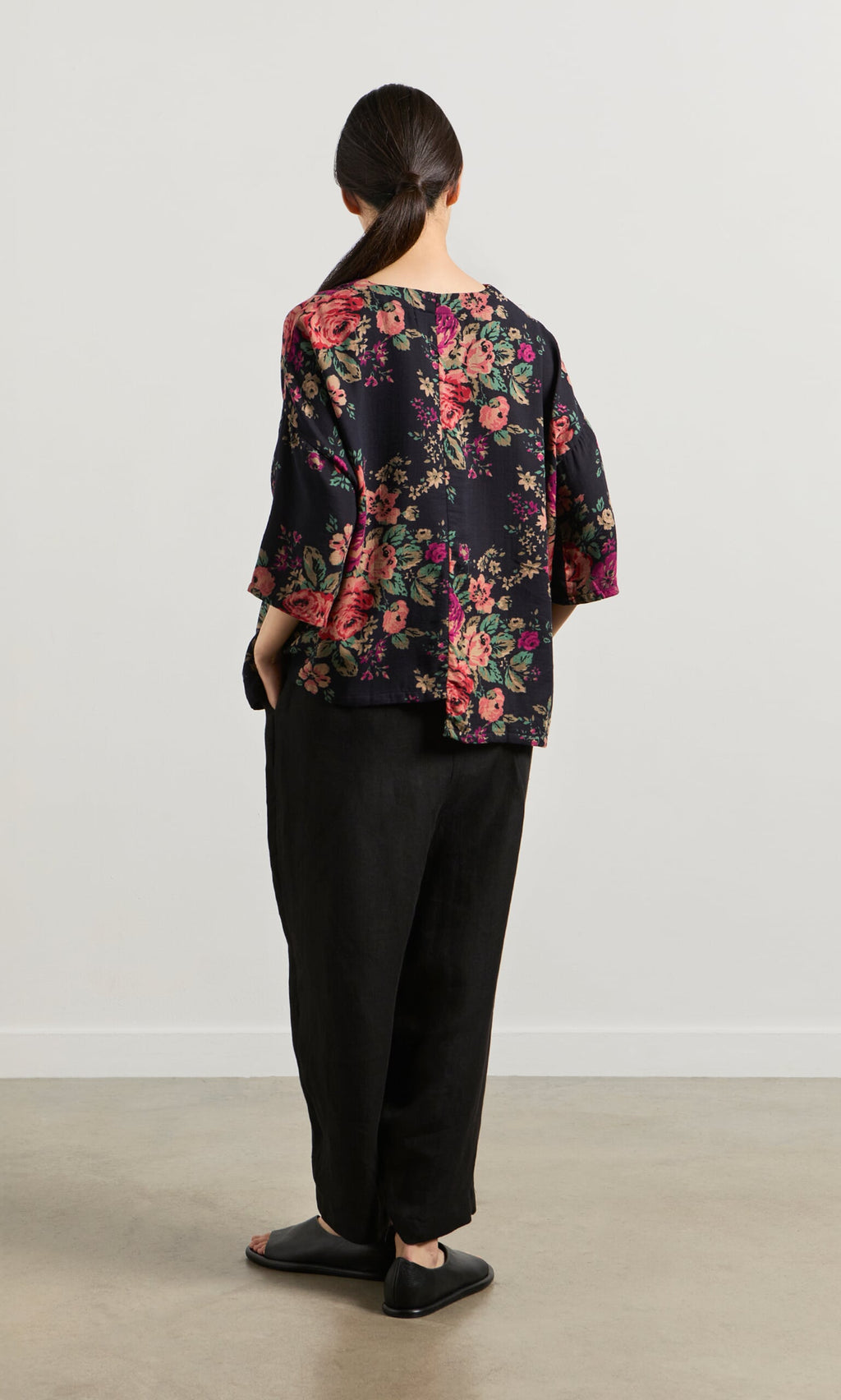 Plümo- Woman with dark hair in a low ponytail wears the Liu Pocket Blouse in Deep Navy, black pants, and sandals, facing away.