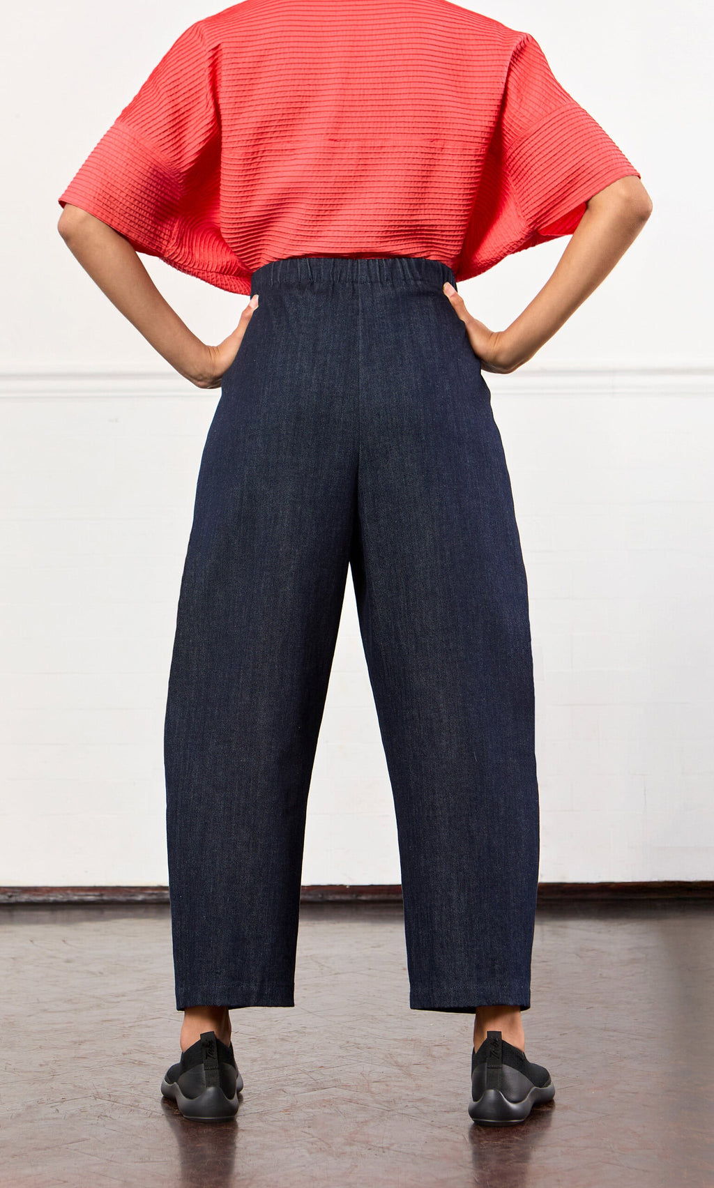 Plümo- Person in a textured red top and Lugo Barrel Jeans, standing with hands on hips and facing away.