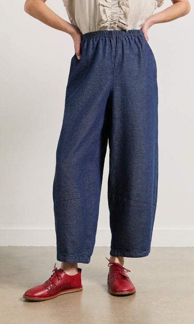 Plümo- Person wearing Mara Barrel Pants in indigo with red shoes, hands in pockets, against a plain background.