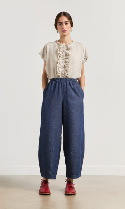 Plümo- Woman stands against a white wall in a beige ruffled top, Mara Barrel Pants in indigo, and red shoes, hands in pockets.