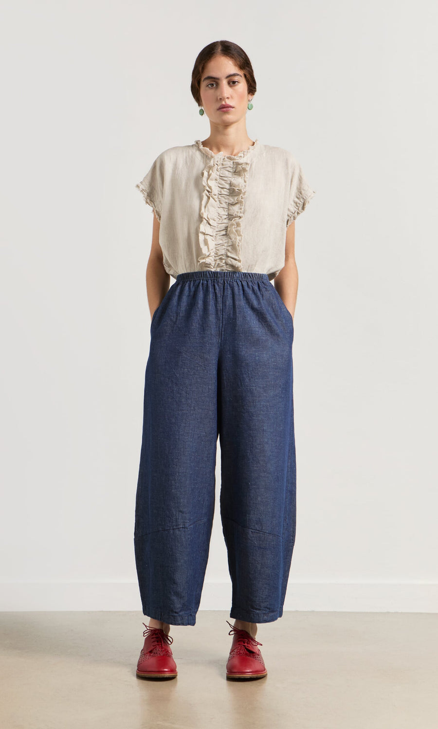 Plümo- Woman stands against a white wall in a beige ruffled top, Mara Barrel Pants in indigo, and red shoes, hands in pockets.