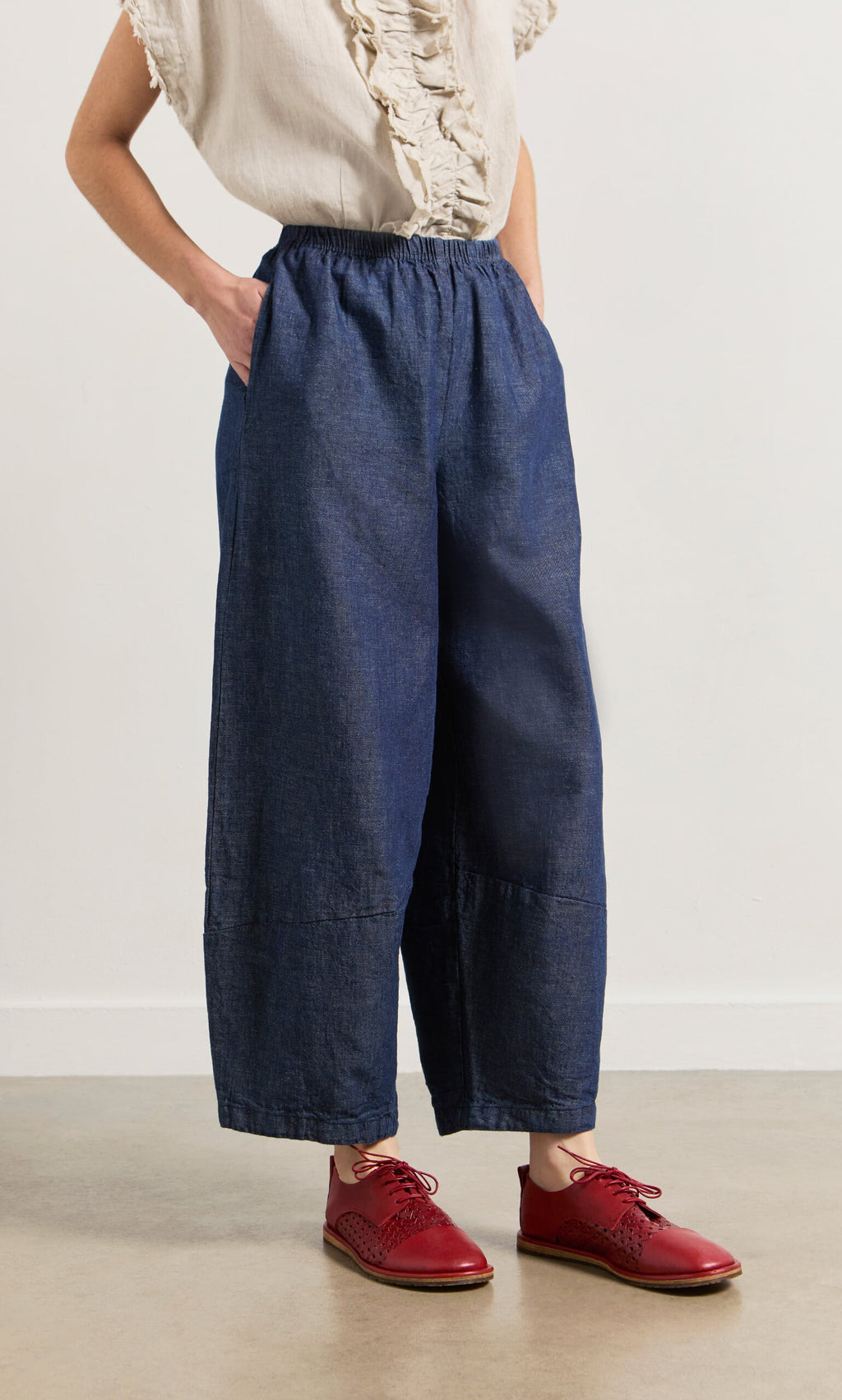 Plümo- Person wearing Mara Barrel Pants in indigo, a cream ruffle top, and red oxford shoes, stands against a plain background.