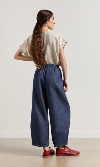 Plümo- A woman faces away in a beige top and Mara Barrel Pants, featuring a dark indigo wash and red shoes.